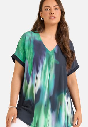 Femme portant un haut ample à col en V avec des manches courtes, présentant un motif abstrait vert, bleu et blanc et des poignets de manches bleu foncé.