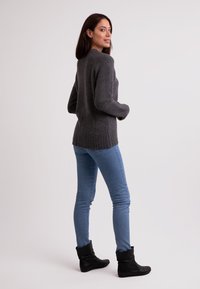 Maglione grigio lavorato a maglia con orlo a coste, indossato sopra jeans skinny blu e stivali neri alla caviglia. Texture liscia con design aderente, visto di lato.