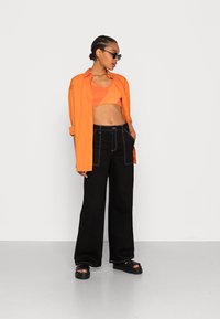 Camisa oversized laranja sobre um top cropped laranja texturizado, combinada com calças largas pretas com costura branca e sandálias pretas.