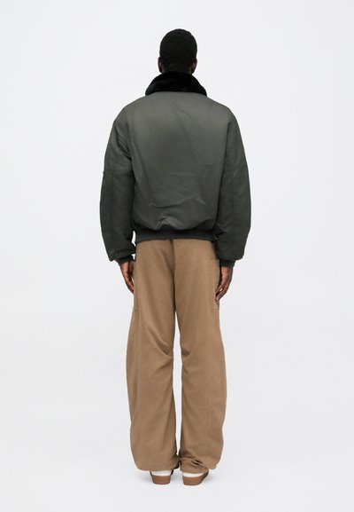 Kohlrabenschwarze Bomberjacke mit einem schwarzen Kunstfellkragen, kombiniert mit beigen weiten Hosen. Glatte Textur, lockere Passform und minimalistisches Design.