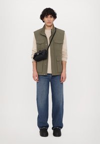 Colete verde-oliva sobre um suéter bege, combinado com jeans de perna larga azuis e sapatos pretos. Uma pequena bolsa cruzada preta pende do ombro.