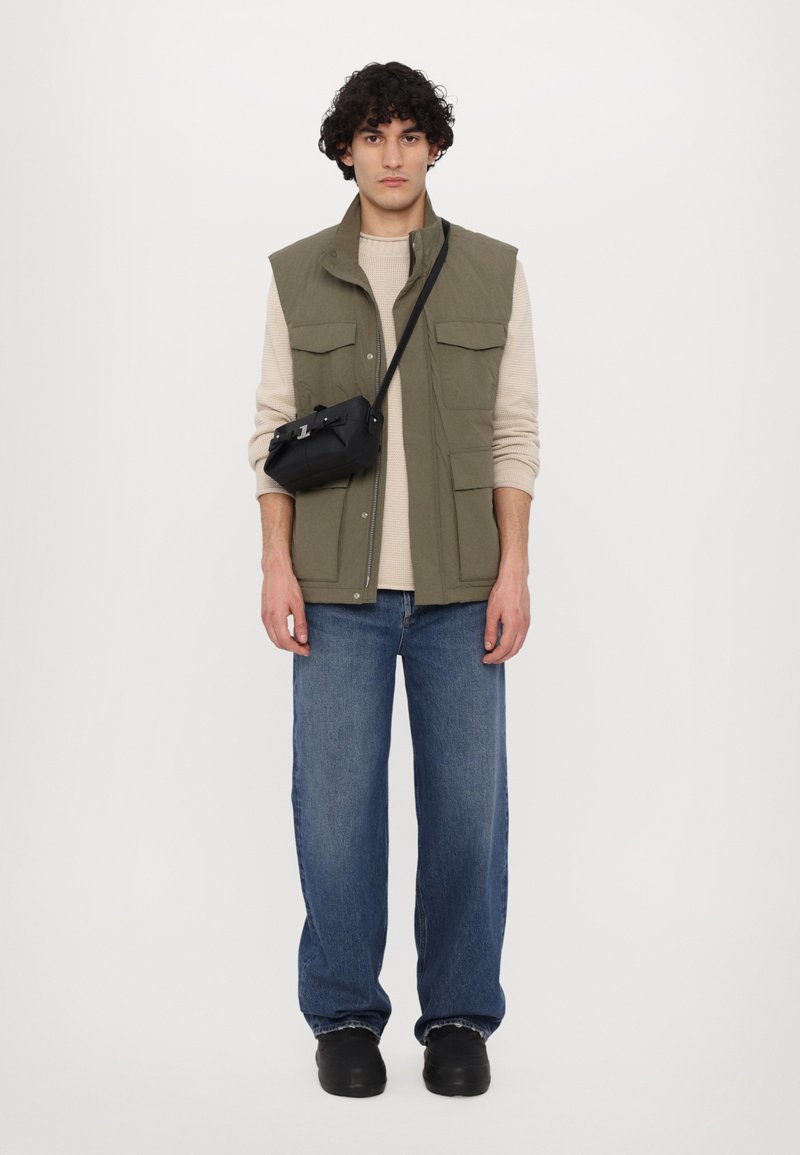 Colete verde-oliva sobre um suéter bege, combinado com jeans de perna larga azuis e sapatos pretos. Uma pequena bolsa cruzada preta pende do ombro.