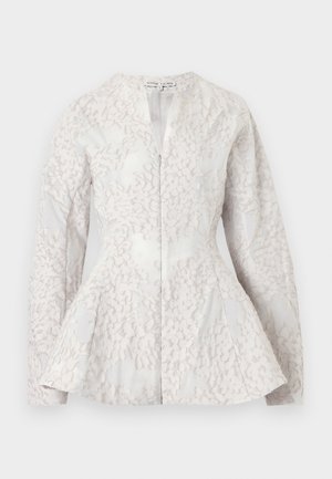 Nainen pukeutuneena teksturoidun kermaisen takin kanssa, jossa on peplum-vyötärö ja yhteen sopiva midi-hame etuhalkiolla, yhdistetty mustiin korkokenkäisiin sandaaleihin.