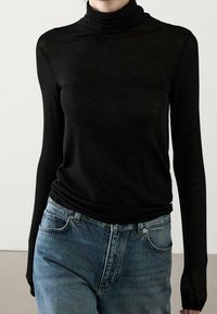 Persoon in een aansluitend zwart hoogsluitend shirt met lange mouwen, gestoken in hoge taille blauwe jeans, tegen een effen achtergrond.