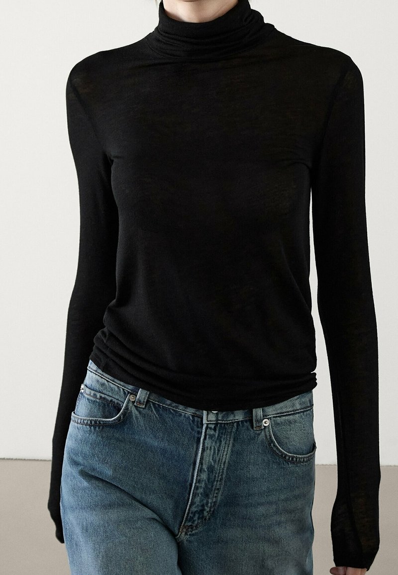 Persoon in een aansluitend zwart hoogsluitend shirt met lange mouwen, gestoken in hoge taille blauwe jeans, tegen een effen achtergrond.