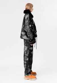 Chaqueta de patrón negro y gris con cuello de piel sintética negra, combinada con pantalones texturizados a juego y zapatos gruesos naranjas.