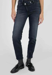 Femme portant un jean slim bleu marine avec une fermeture à double bouton superposée, des chaussettes grises et des mocassins noirs brillants, se tenant devant un fond uni.