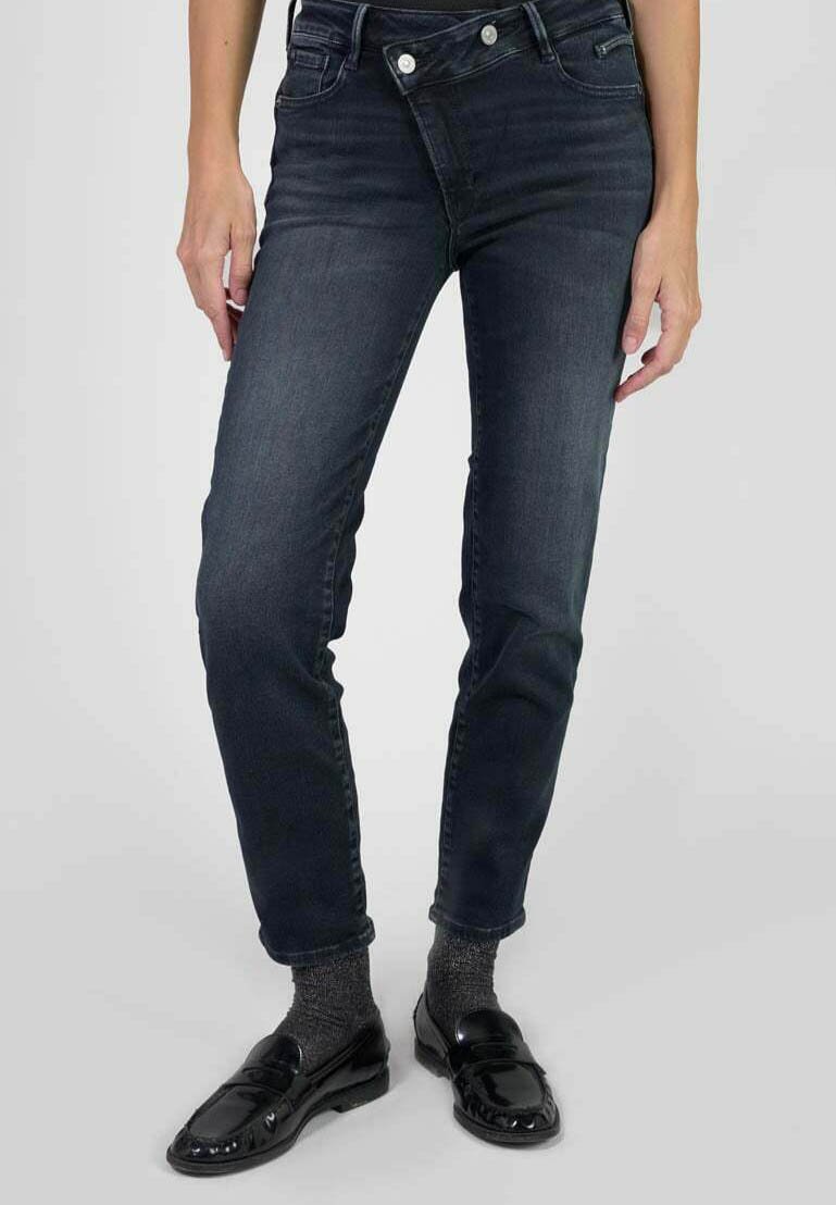 Femme portant un jean slim bleu marine avec une fermeture à double bouton superposée, des chaussettes grises et des mocassins noirs brillants, se tenant devant un fond uni.