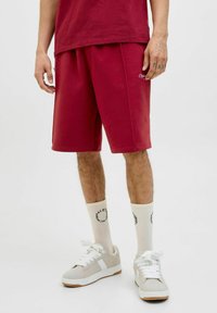 Shorts de sport bordeaux avec une taille élastique, poches latérales et une coupe ample, associés à des baskets gris clair et des chaussettes crème avec un logo.