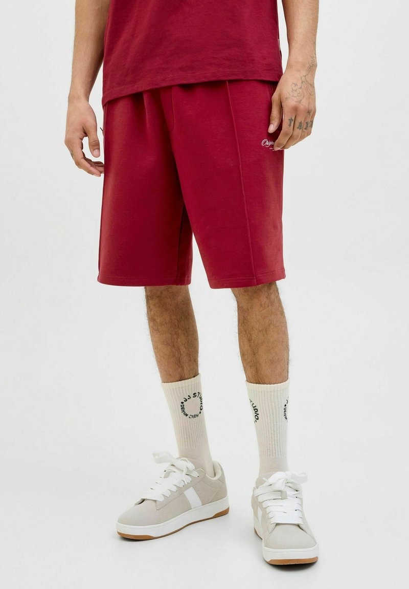 Shorts de sport bordeaux avec une taille élastique, poches latérales et une coupe ample, associés à des baskets gris clair et des chaussettes crème avec un logo.