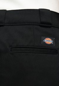 Κοντινό πλάνο της πίσω τσέπης μαύρου παντελονιού Dickies με λογότυπο και θηλιές για ζώνη από πάνω, φορεμένο με λευκό ύφασμα ορατό στην κορυφή.