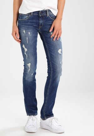 Woman wearing slim-fit blue distressed jeans, white sneakers, and a white top, standing against a plain light background.