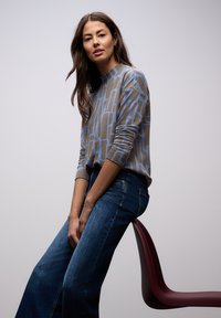 Jersey marrón claro con patrón geométrico azul, combinado con vaqueros anchos azul oscuro. Modelo sentado en una silla granate. Fondo neutro.