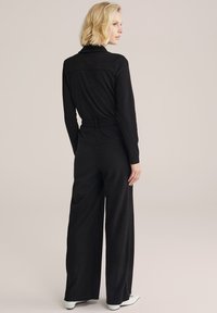 Melna platā kājā jumpsuits ar garām piedurknēm un jostu ap vidu. Tekstūrēta auduma. Modelis ir noraidījis uz sāniem ar plecu garuma matiem.