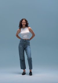 Valge keha jälgiv topp, mis on sobitatud hele siniste kõrge vöökohaga teksadega. Modell kannab musti kingi; taust on ühtlane helesinine.