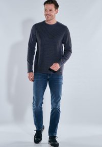 Dunkelgraues Langarmshirt mit strukturiertem Design, kombiniert mit blauen Jeans und schwarzen Sneakers. Modell steht aufrecht vor einem neutralen Hintergrund.