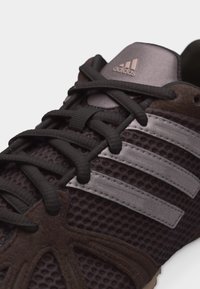 Σκούρο καφέ αθλητικό παπούτσι Adidas με μαύρα κορδόνια, επάνω μέρος από δίχτυ, σουέντ λεπτομέρειες και τρεις αντανακλαστικές ασημί ρίγες στο πλάι.