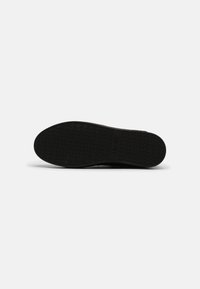 Suela de zapato de goma negra plana con una superficie texturizada, que presenta patrones circulares y un logotipo en relieve en la zona de la mediasuela.