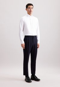 Camicia bianca a maniche lunghe con colletto abbottonato, abbinata a pantaloni eleganti blu navy e scarpe in pelle nera. Tessuto morbido e design aderente.