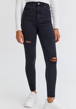 Woman wearing high-waist black skinny jeans with ripped knees, white long-sleeve top, and white sneakers against plain background.