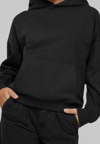 Persona vestida con una sudadera negra y pantalones negros, con las manos en los bolsillos, mostrada desde la mitad del pecho hasta las caderas contra un fondo liso.