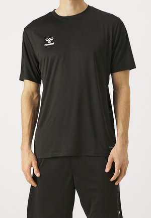 Mies pukeutuneena yksinkertaiseen mustaan lyhythihaiseen urheilupaitaan, jossa valkoinen Hummel-logo rinnassa, ja mustiin yhteensopiviin shortseihin.