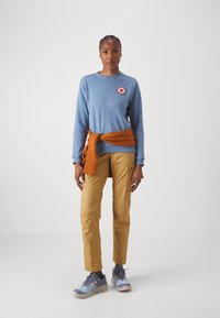 Blaues Sweatshirt mit Logo, orangefarbener Pullover um die Taille gebunden, beiger Cargo-Hose und graue Sneaker mit rosa Akzenten.