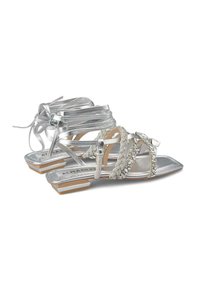Sandales métalliques argentées avec des brides décoratives, dotées d'un talon plat et de détails ornés. Texture lisse et design à lacets.