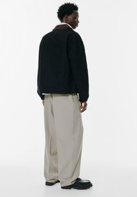 Schwarze übergroße Jacke mit braunem Kragen, kombiniert mit hellbeigen, weit geschnittenen Hosen und schwarzen Schuhen. Glattes Material und lockerer Schnitt.