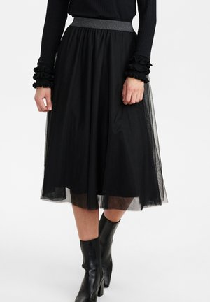 Femme portant une jupe midi noire avec un voile transparent, un haut noir avec des poignets à volants et des bottines noires à talons.