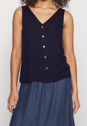 Kobieta ubrana w granatowy, bezrękawnik z guzikami z przodu i dekoltem w serek, zestawiona z wysokim stanem niebieskiej spódnicy dżinsowej, dłonie swobodnie ułożone wzdłuż boków, neutralne tło.