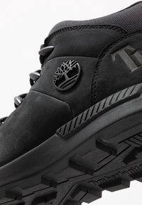 Gros plan d'une chaussure de randonnée Timberland noire montrant une tige en cuir, des œillets métalliques, le logo en forme d'arbre et une semelle en caoutchouc texturée pour une meilleure adhérence.
