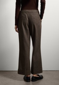 Bruine tweed broek met wijde pijpen en een getailleerde pasvorm, twee achterzakken, omgeslagen pijpen en gedragen met zwarte loafers.