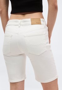 Γυναίκα που φοράει λευκά τζιν σορτς μέχρι το γόνατο με πίσω τσέπες και ταμπελάκι μάρκας σε καφέ χρώμα, συνδυασμένα με μαύρο κοντό τοπ.