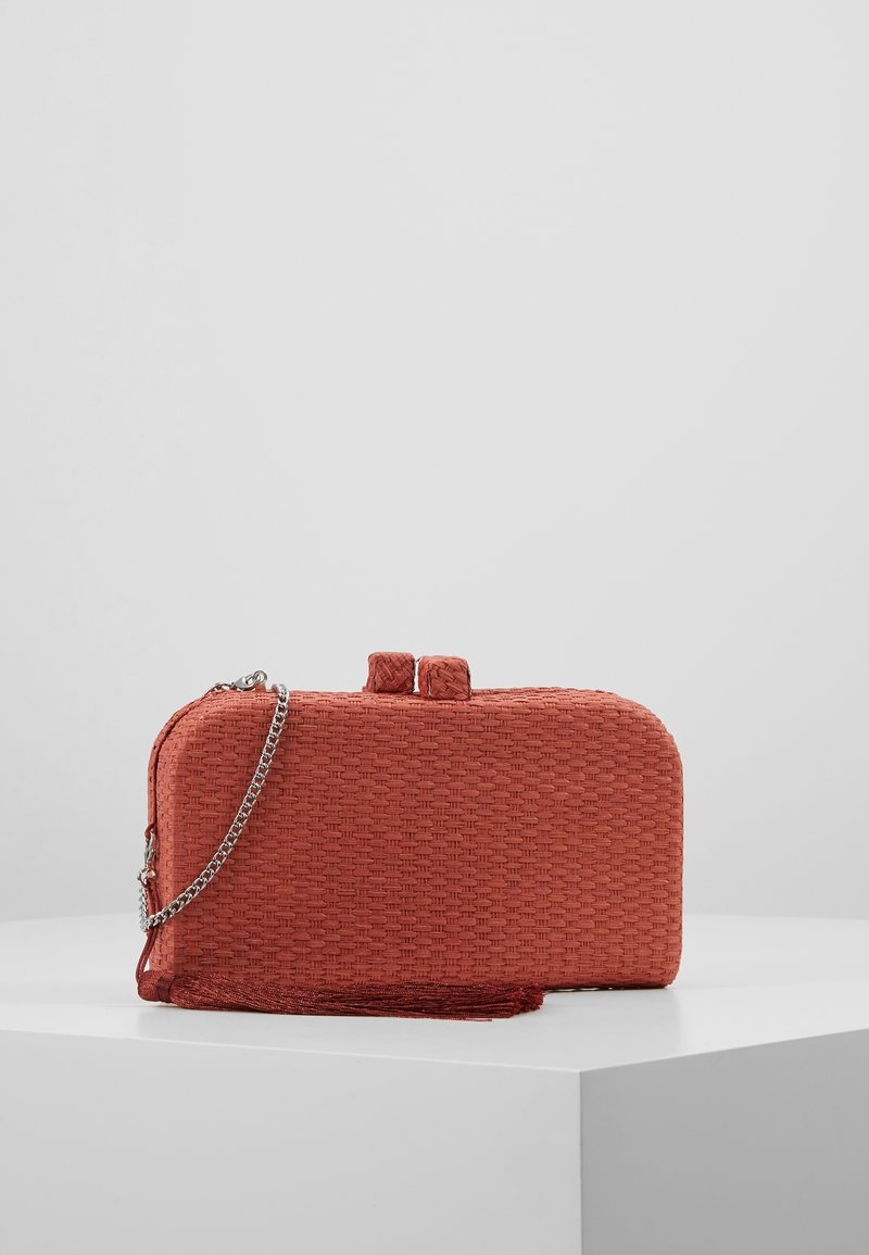 Petit clutch rectangulaire tissé couleur rouille-orange avec bandoulière chaîne argentée, pompon en tissu et double fermeture arrondie sur surface blanche.