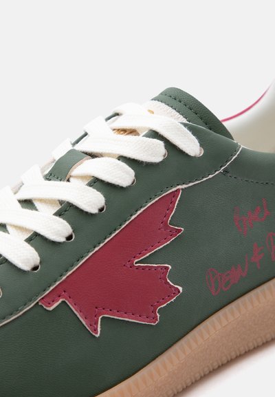 Πράσινο αθλητικό παπούτσι με λευκά κορδόνια, ένα ραμμένο κόκκινο φύλλο σφένδαμου στην πλευρά, κόκκινο χειρόγραφο κείμενο και ανοιχτό καφέ σόλα.