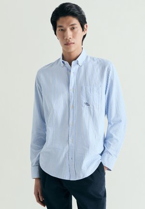 Joven con camisa de botones a rayas verticales en azul claro y blanco, con un bolsillo en el pecho, combinada con pantalones oscuros, de pie frente a un fondo liso.