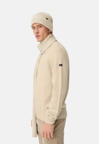 Beige gerippter Strickpullover mit passendem Schal und Mütze. Verfügt über ein figurbetontes Design und ein kleines Logo-Patch am Ärmel. Weicher Stoff.