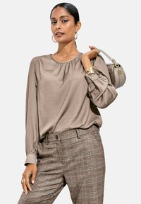 Langärmlige taupe Bluse mit rundem Ausschnitt und gefälteltem Detail, kombiniert mit braunen karierter Hose. Accessoires mit Ohrringen und Handtasche.
