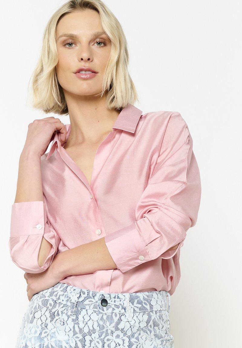 Roze satijnen shirt met een kraag en knoopmanchetten, met een gladde textuur; gecombineerd met witte broek met kantpatroon.