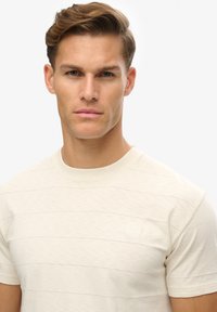 Camiseta color beige claro con sutiles rayas horizontales, cuello redondo y un pequeño logo en relieve en el pecho, hecha de una tela suave.