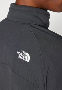 Šedý fleecový pulovr s vysokým límcem, zdobený mřížkovaným vzorem a bílým logem "The North Face" na levém rameni.