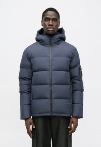 Tengerészeti kék puffermellény, cipzárral az elülső részen, kapucnival és látható varrással. Oldalzsebekkel és szövetpólóval a bal karon rendelkezik. Simaszerkezetű.