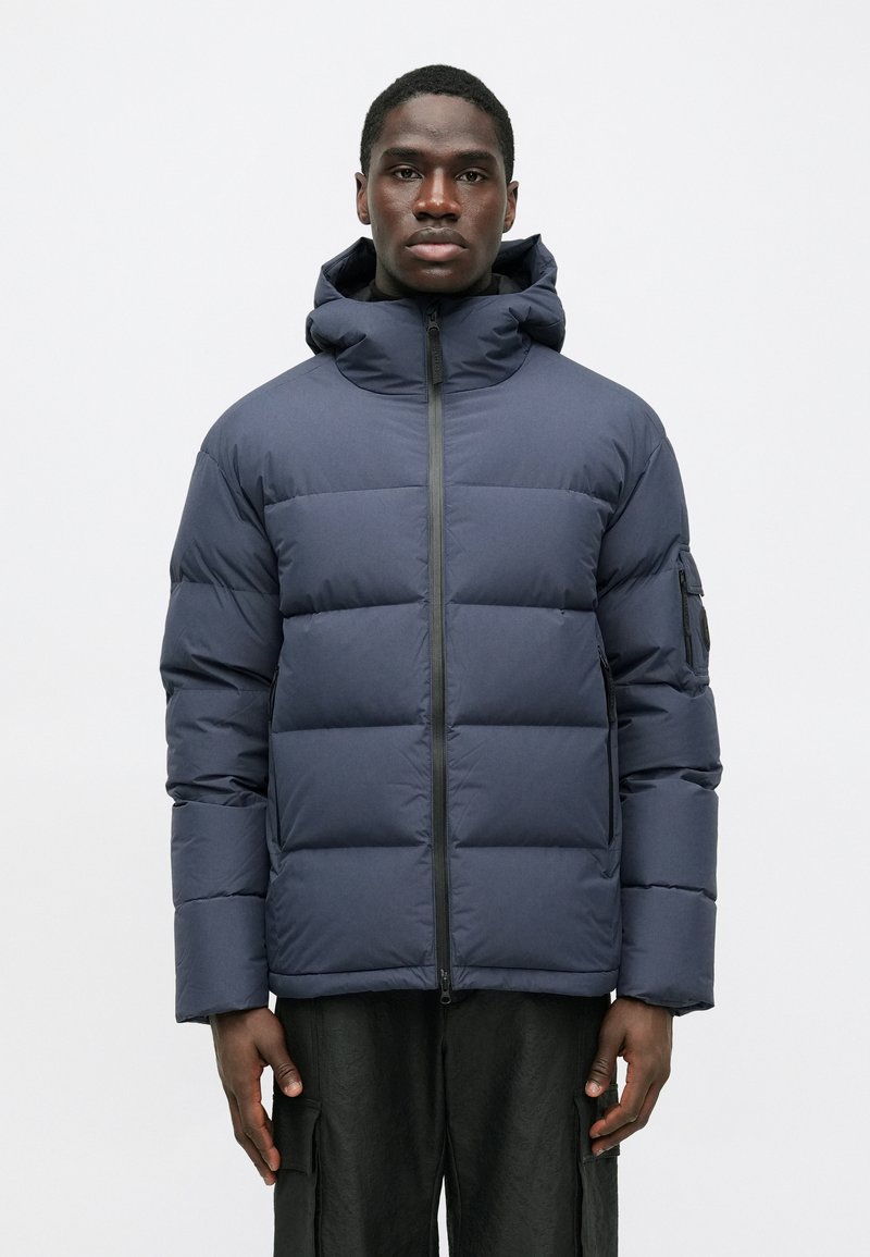 Tengerészeti kék puffermellény, cipzárral az elülső részen, kapucnival és látható varrással. Oldalzsebekkel és szövetpólóval a bal karon rendelkezik. Simaszerkezetű.