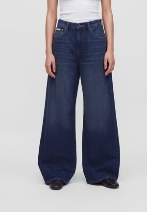Personne portant un jean bleu foncé à jambes larges et des chaussures noires, debout les bras détendus le long du corps devant un fond uni.