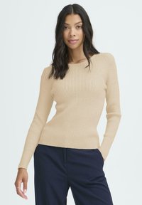 Giovane donna con lunghi capelli scuri, vestita con un maglione in cotone beige a coste e pantaloni blu navy, in piedi con una mano in tasca.