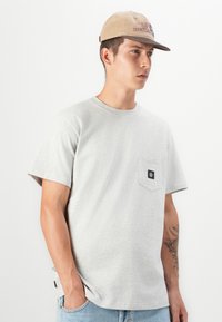 Gray cotton t-shirt with short sleeves, featuring a left chest pocket and a small logo patch. Paired with light blue denim jeans.