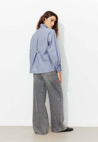 Chemise rayée purple et verte avec un col haut et une fente au dos, associée à un large jean gris. Des chaussures noires à studs complètent le look.