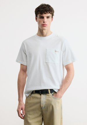 Tânăr purtând un tricou alb cu guler rotund și buzunar la piept și pantaloni bej, stând cu o mână în buzunar pe un fundal simplu.