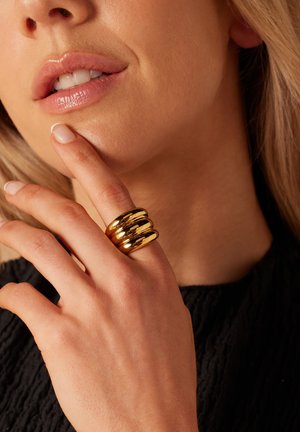 Goldring mit drei geschwungenen Bändern. Glatte, glänzende Oberfläche. Die Hand des Models ruht auf dem Kinn, die Nägel sind lackiert, die Haut hat einen natürlichen Hautton und eine natürliche Textur.